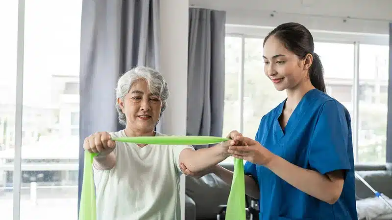 Physiotherapist helping an older woman do resistance band exercises during post-operative rehabilitation in London